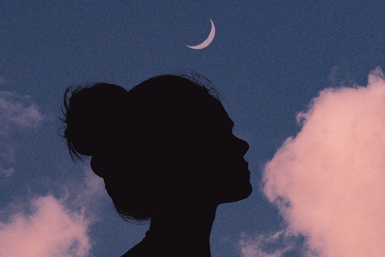 Silhouette nera di una ragazza di profilo con capelli raccolti in uno chignon. Sullo sfondo, un cielo azzurro, nuvole rosa e la luna crescente in lontananza